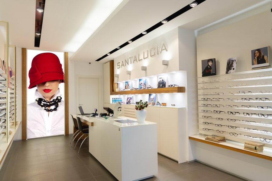 L'interno di Ottica Santalucia: le pareti di occhiali, il banco bianco e il pannello luminoso col cappello rosso