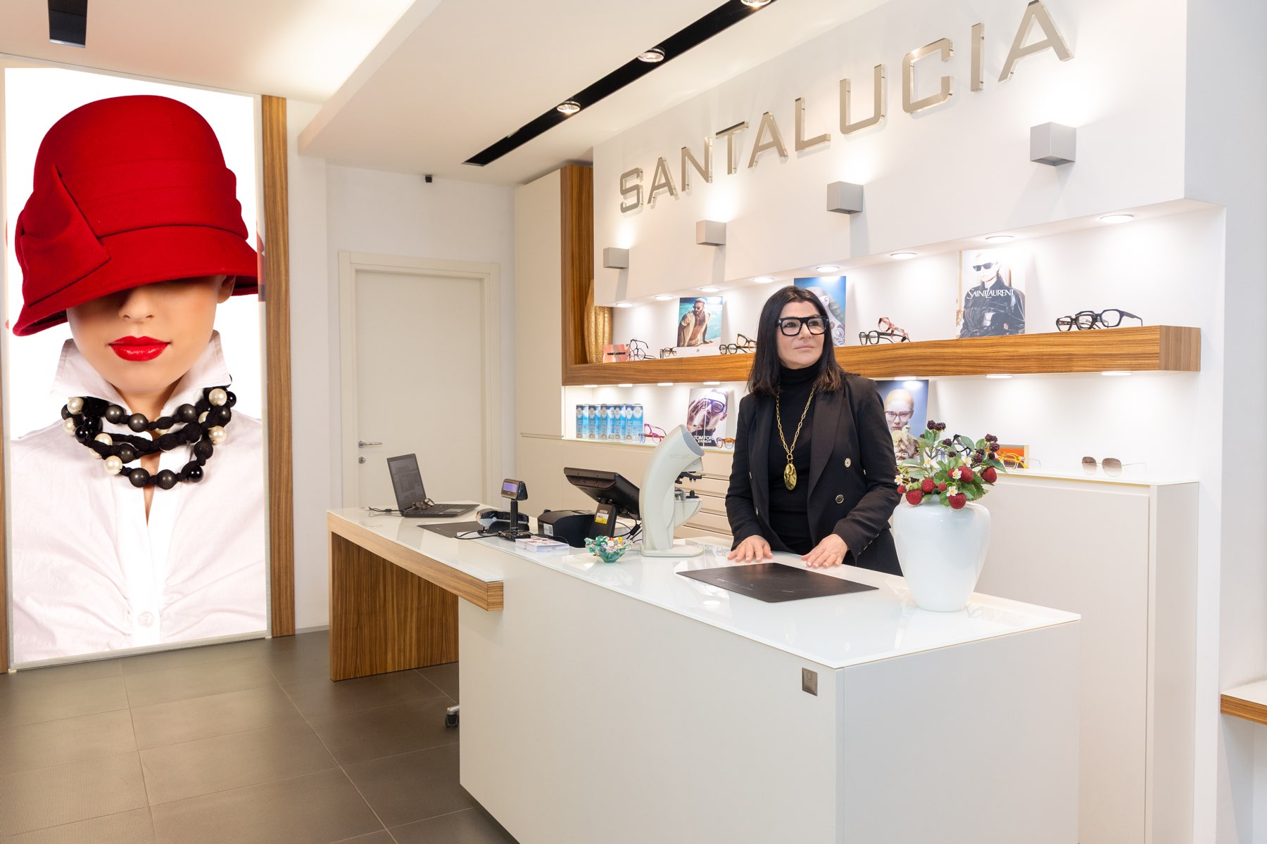 Il negozio Ottica Santalucia a Porto Garibaldi, con Elisa al banco e il pannello luminoso della donna col cappello rosso
